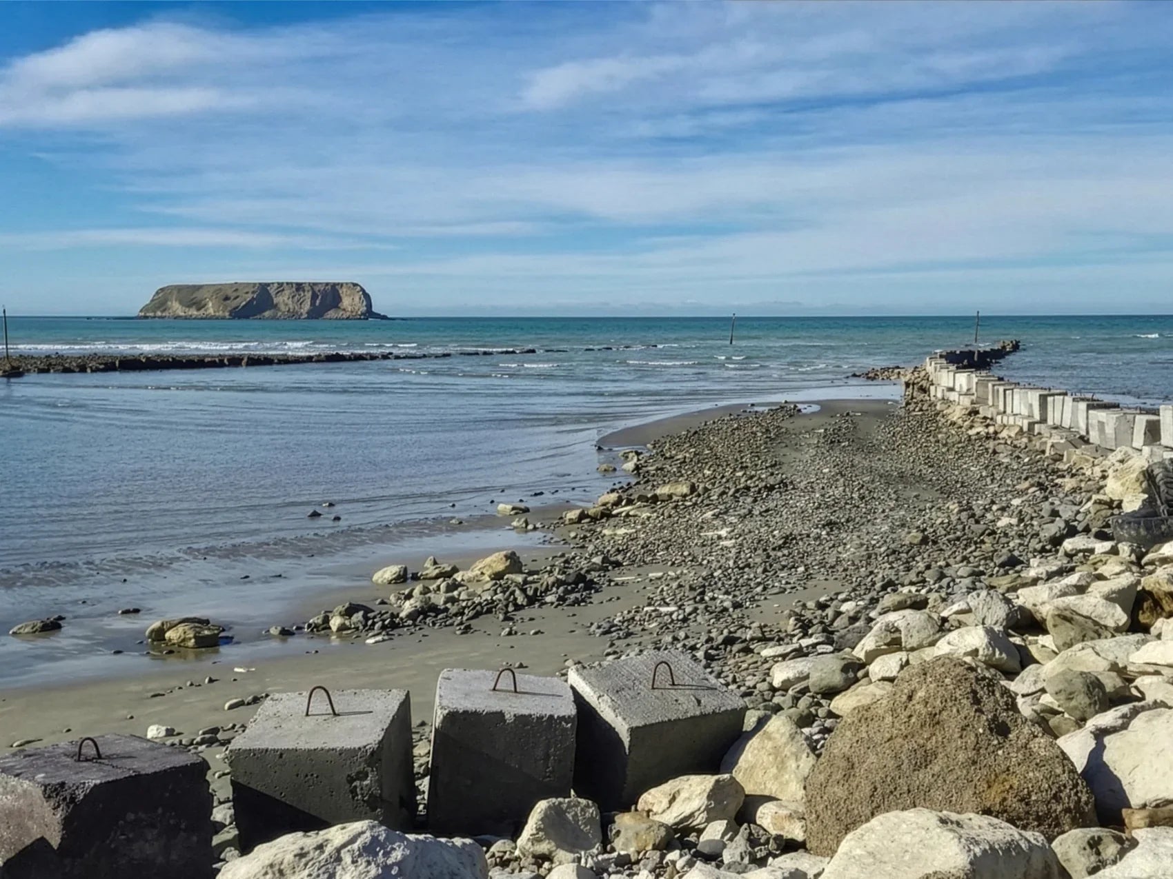 A row of large concrete blocks forming a protective sea wall along a rocky shoreline, with a small island visible on the horizon under a clear blue sky.